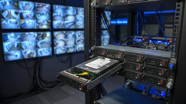 An open rackmount surveillance server with a row of hard drives, one drive pulled partway out, in a server room with monitors showing camera grids
